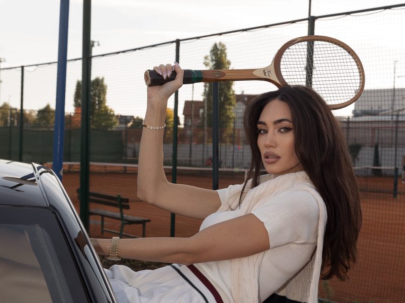 Dubravka Švegar in white tennis clothes with a wooden racket, by a car next to a tennis court